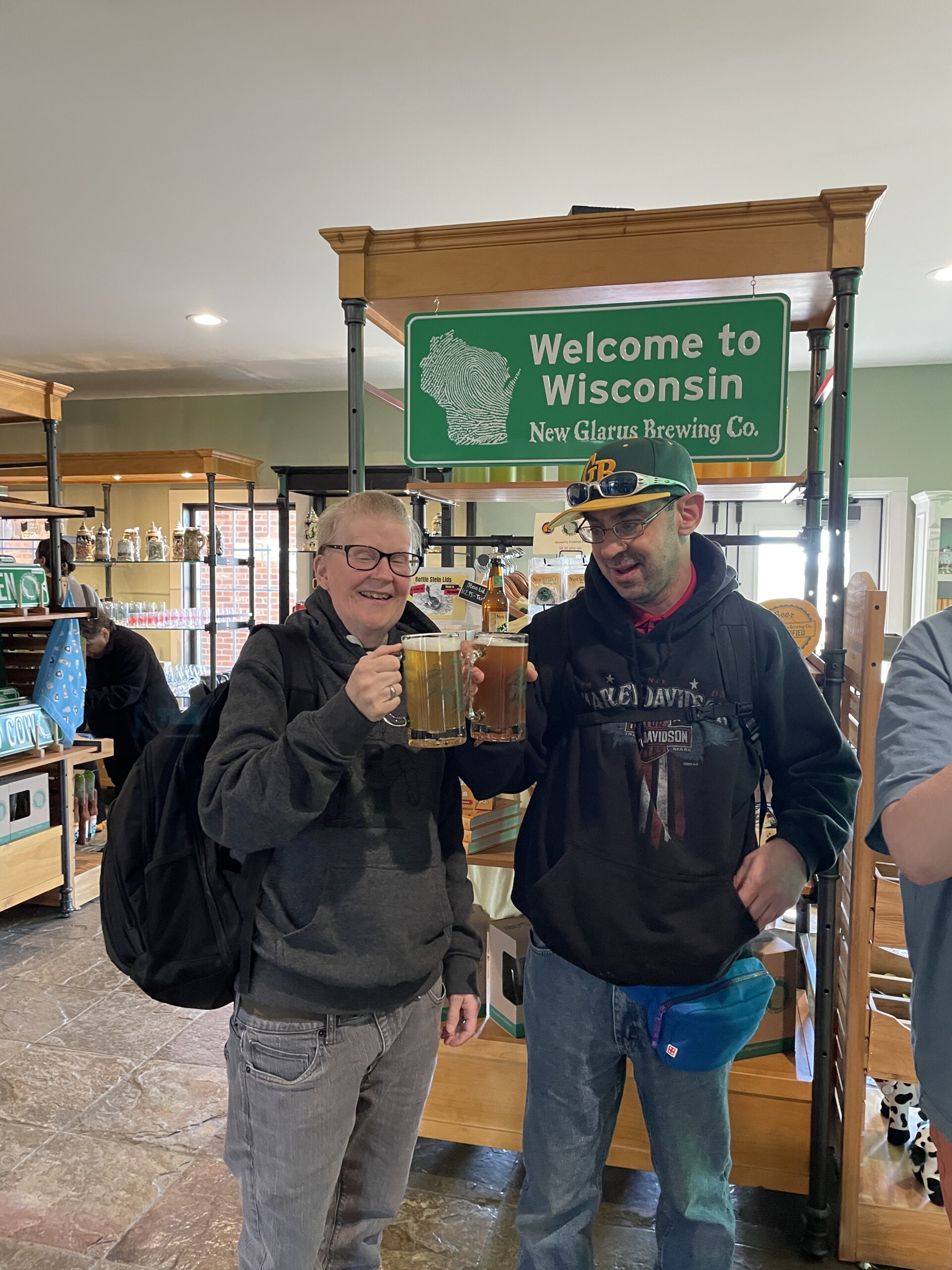 A picture of two male CEL members toasting beers in front of a 'Welcome to Wisconsin' sign.