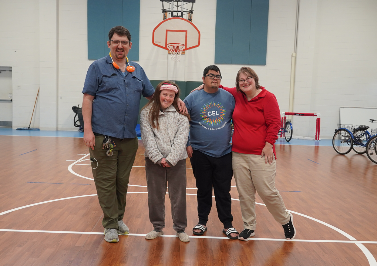 Smiling Catalyst life skills members with two staff members. One member wears a burgundy long sleeve shirt and holds a piece of paper, another member is in a blue jacket, and a third member has a blue shirt. One staff member is in a grey top, and the other in a white graphic tee.