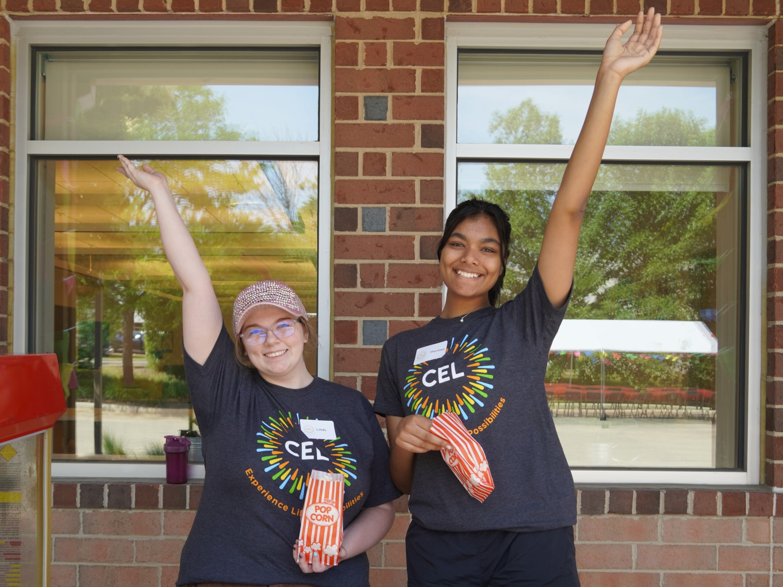 CEL staff smiling during the community day event, with both hands raised, holding popcorn bags wearing grey CEL Merch shirt.