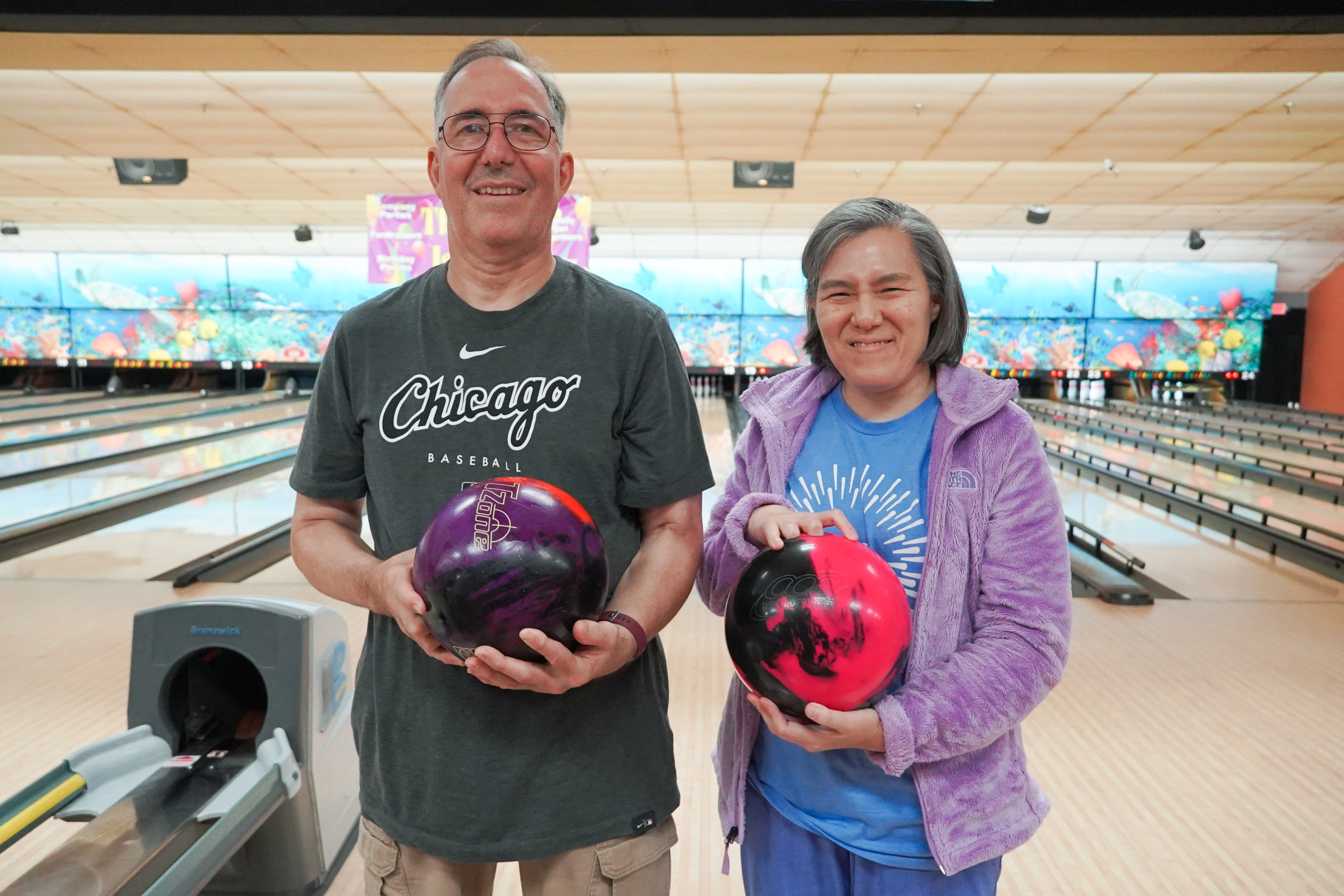 Two members smile and hold bowling balls at a bowling alley